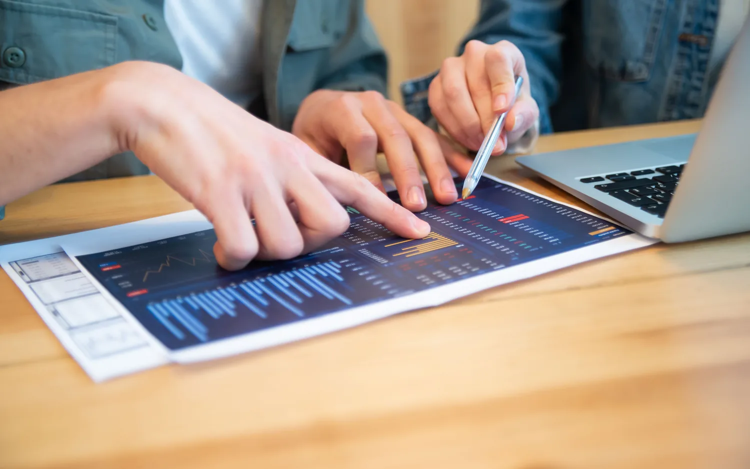 Duas pessoas analisam um painel financeiro impresso sobre a mesa, ao lado de um notebook.