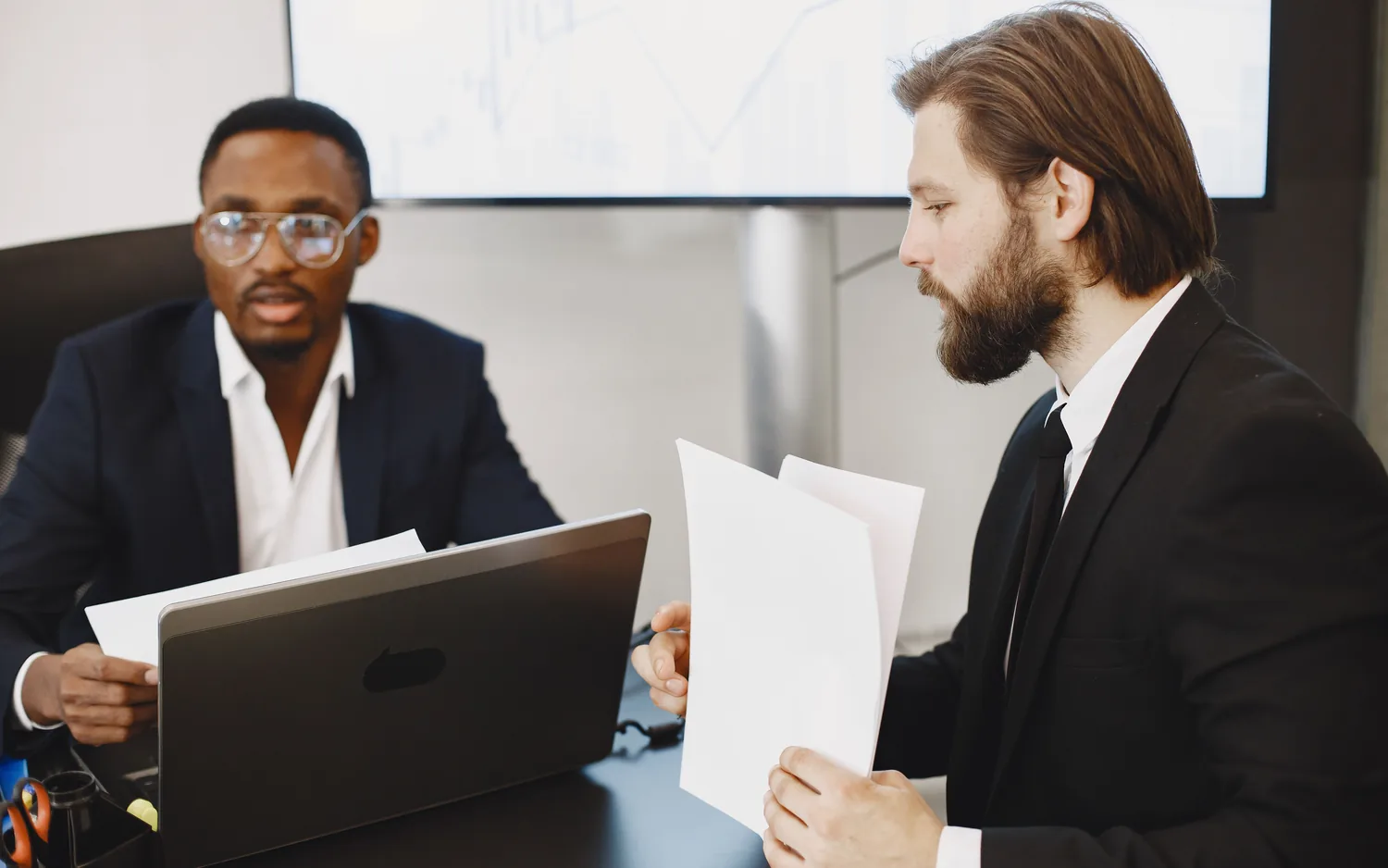Dois executivos conversam à mesa enquanto um deles compara duas folhas de papel.